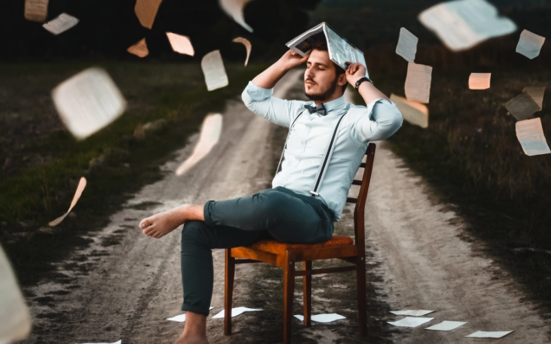 A man sitting in a chair, holding a book open on his head. He is not wearing socks, and is positioned on a remote dirt road. Many papers fall from the sky around him.