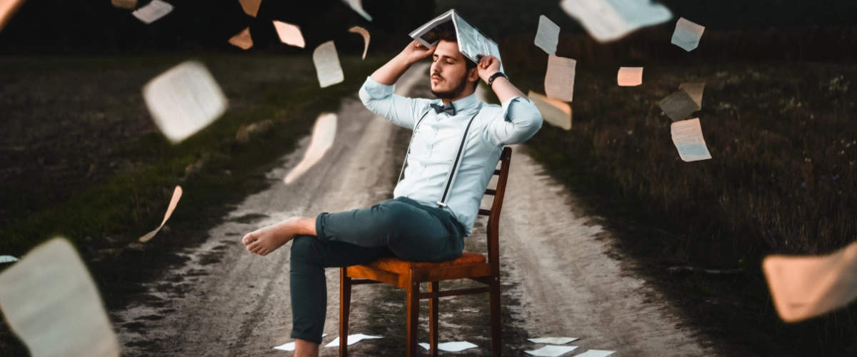 A man sitting in a chair, holding a book open on his head. He is not wearing socks, and is positioned on a remote dirt road. Many papers fall from the sky around him.