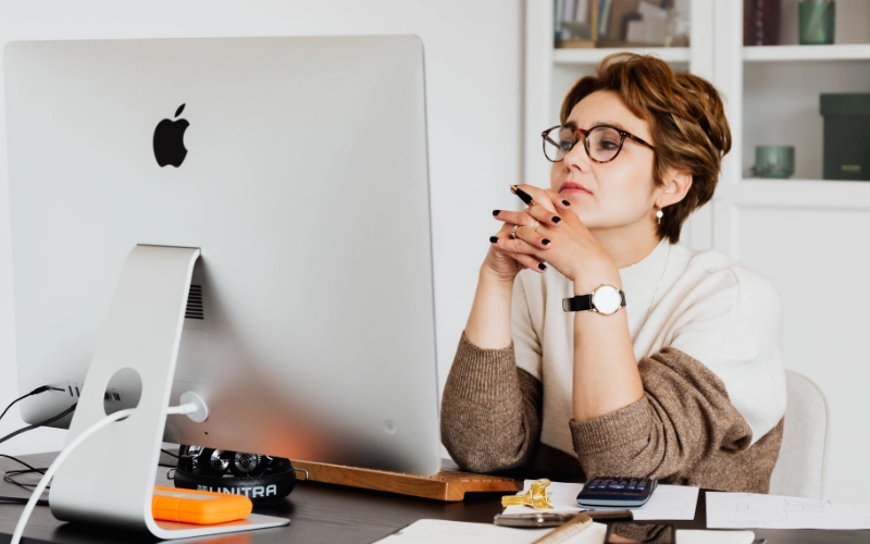 A woman sitting at a desk, looking at her computer, and thinking.