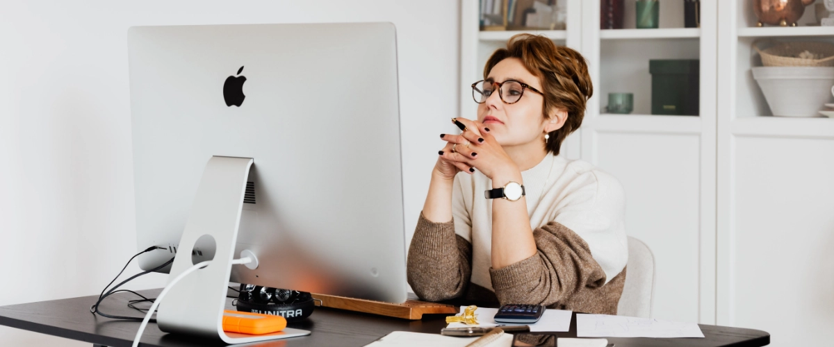 A woman sitting at a desk, looking at her computer, and thinking.